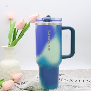 Puede incluir: Un vaso grande y aislado con asa. El vaso es principalmente azul con un degradado de azul claro y verde. La marca "STANLEY" est&aacute; impresa en el lateral. Tiene una tapa transparente y est&aacute; dise&ntilde;ado para bebidas.