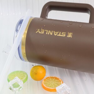 May include: A brown Stanley thermos with a handle and a gold band. The thermos is wet with water droplets. The image also includes a lime slice, an orange slice, a whole orange, and ice cubes.