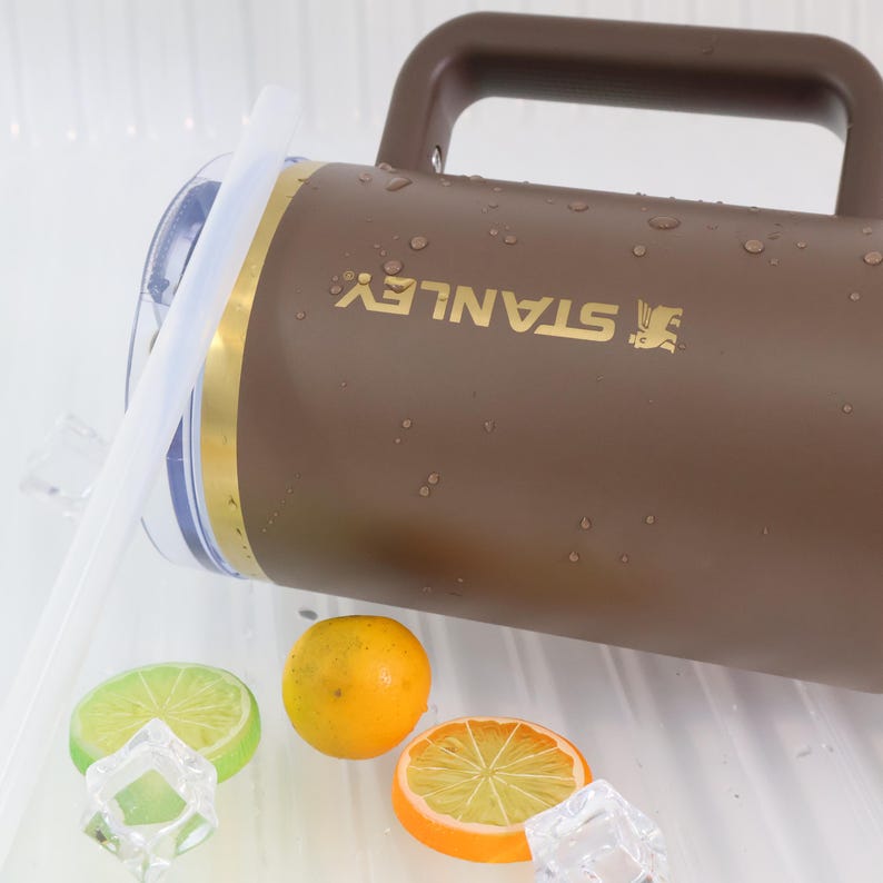 May include: A brown Stanley thermos with a handle and a gold band. The thermos is wet with water droplets. The image also includes a lime slice, an orange slice, a whole orange, and ice cubes.