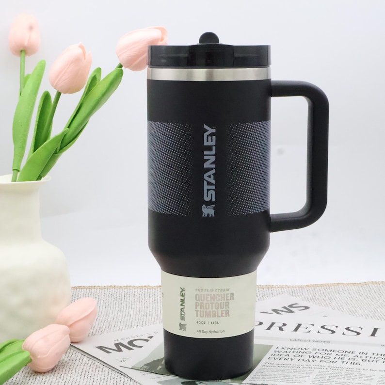 Puede incluir: Vaso Stanley Quencher Protour negro con asa y tapa negra. El vaso tiene una banda texturizada y una etiqueta blanca con informaci&oacute;n del producto. El vaso es de 1,18L.