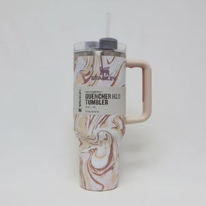 K&ouml;nnte beinhalten: Ein Stanley Quencher H2.0 Tumbler mit Griff und Strohhalm. Der Becher hat ein Wirbelmuster in Beige, Braun und Wei&szlig;. Das Etikett lautet "The Flowstate Quencher H2.0 Tumbler" und hat das Stanley-Logo. Entwickelt f&uuml;r die ganzt&auml;gige Fl&uuml;ssigkeitszufuhr.