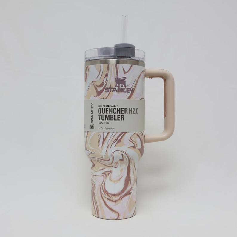 K&ouml;nnte beinhalten: Ein Stanley Quencher H2.0 Tumbler mit Griff und Strohhalm. Der Becher hat ein Wirbelmuster in Beige, Braun und Wei&szlig;. Das Etikett lautet "The Flowstate Quencher H2.0 Tumbler" und hat das Stanley-Logo. Entwickelt f&uuml;r die ganzt&auml;gige Fl&uuml;ssigkeitszufuhr.