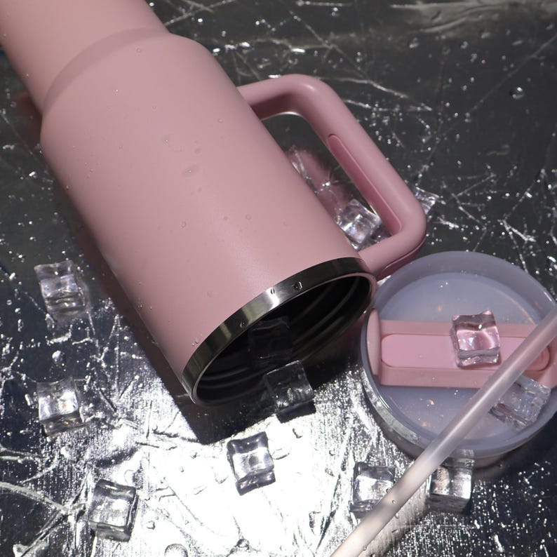 K&ouml;nnte beinhalten: Ein rosa isolierter Becher mit Griff und silbernem Rand. Der Deckel ist abgenommen, wodurch das Innere sichtbar wird, und ein klarer Strohhalm ist zu sehen. Eisw&uuml;rfel sind auf einer reflektierenden Oberfl&auml;che um den Becher verstreut.