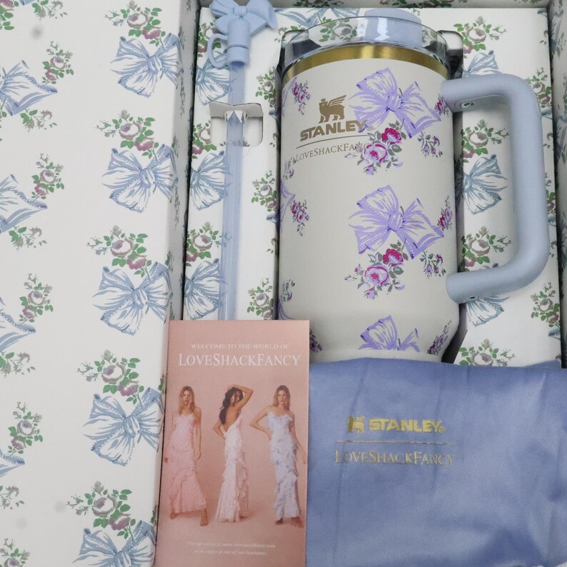 May include: A white Stanley mug with a light blue handle and lid, adorned with purple bows and floral patterns. The mug is packaged in a box with matching floral designs and includes a straw and a brochure with the text "LOVESHACKFANCY".