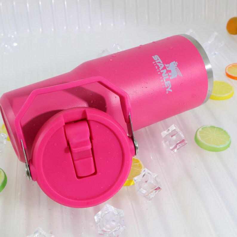 May include: A pink Stanley tumbler with a handle and a flip-top lid. The tumbler has the Stanley logo printed on the side. The tumbler is surrounded by ice cubes and lemon slices.