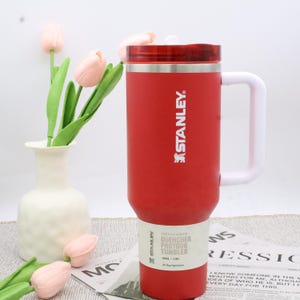 Pu&ograve; includere: Un bicchiere Stanley Quencher Protour rosso con manico e coperchio bianchi. Il bicchiere ha il logo Stanley e le informazioni sul prodotto stampate. Il bicchiere &egrave; da 1,18L. Un vaso bianco con tulipani rosa &egrave; sullo sfondo.