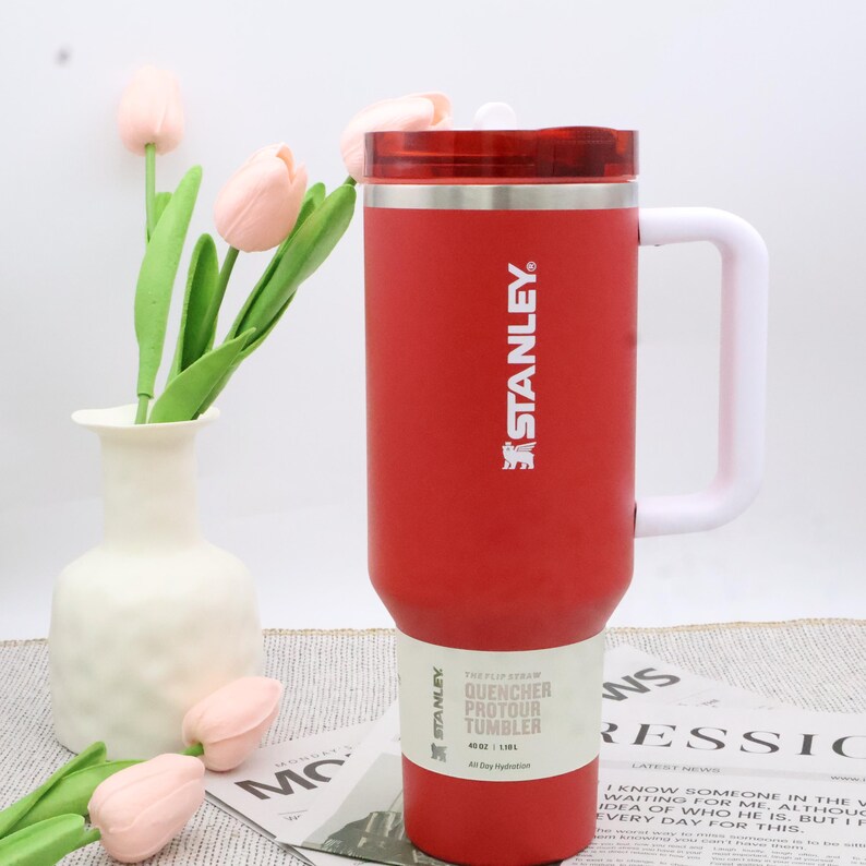 Pu&ograve; includere: Un bicchiere Stanley Quencher Protour rosso con manico e coperchio bianchi. Il bicchiere ha il logo Stanley e le informazioni sul prodotto stampate. Il bicchiere &egrave; da 1,18L. Un vaso bianco con tulipani rosa &egrave; sullo sfondo.