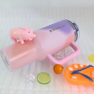 Puede incluir: Un vaso Stanley rosa y morado con asa, con el logotipo de Stanley. Una peque&ntilde;a figura de cerdo rosa se encuentra encima. El vaso est&aacute; rodeado de cubitos de hielo, una rodaja de lima, una naranja y una chancla naranja.
