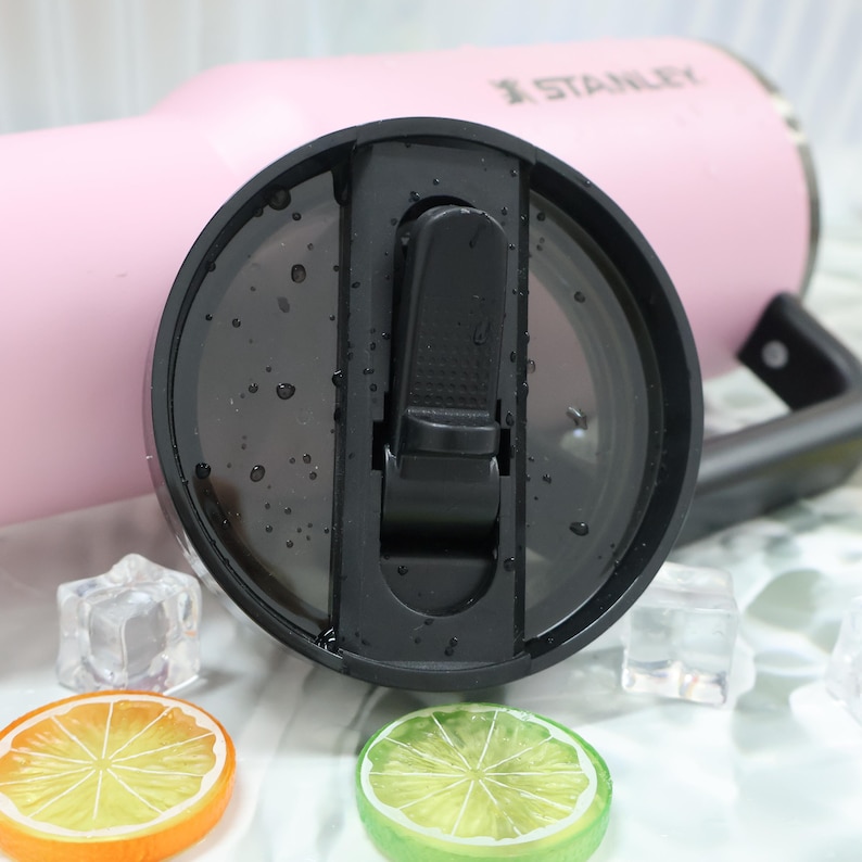 May include: Close-up of a pink Stanley tumbler with a black lid. The lid is open, revealing the interior. The tumbler has a black handle. Ice cubes and citrus fruit slices are in the foreground.