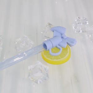 K&ouml;nnte beinhalten: Ein hellblauer Getr&auml;nkespenderhahn mit Schleifendetail, der auf einer gelben Zitronenscheibe ruht. Ein klarer Schlauch erstreckt sich vom Wasserhahn. Klare Eisw&uuml;rfel sind um den Wasserhahn und die Zitronenscheibe verstreut.