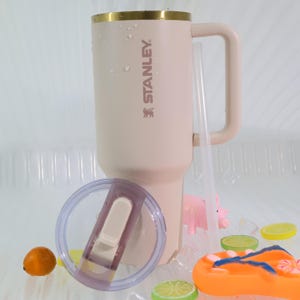 Puede incluir: Un vaso Stanley grande, de color rosa claro, con borde dorado, asa y el nombre de la marca impreso en el lateral. Se incluye una pajita y una tapa transparente. El vaso est&aacute; rodeado de hielo, rodajas de fruta y una peque&ntilde;a figura de cerdo.