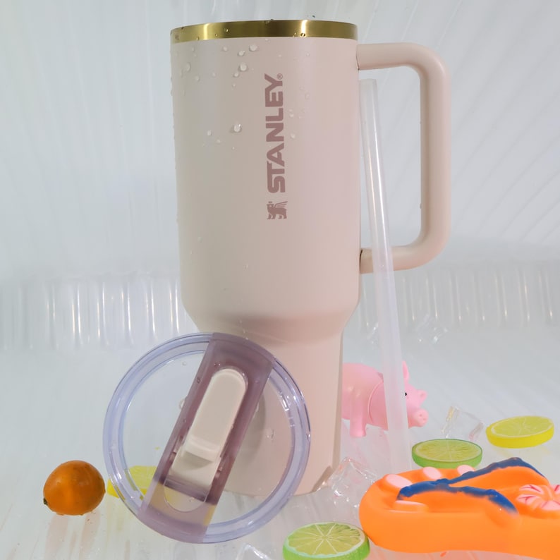 Puede incluir: Un vaso Stanley grande, de color rosa claro, con borde dorado, asa y el nombre de la marca impreso en el lateral. Se incluye una pajita y una tapa transparente. El vaso est&aacute; rodeado de hielo, rodajas de fruta y una peque&ntilde;a figura de cerdo.