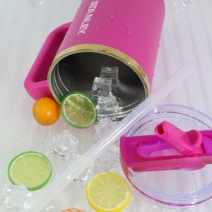 May include: A pink Stanley tumbler with a handle, gold trim, and the brand name visible. The tumbler is open, revealing ice cubes inside. A clear straw and lid are nearby, along with slices of lime, lemon, and an orange.