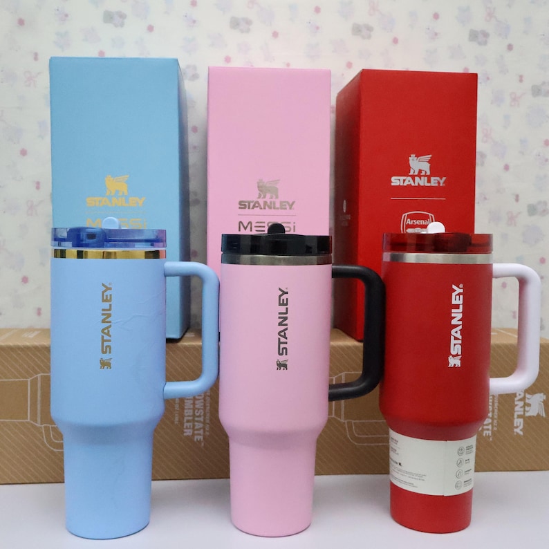 Pu&ograve; includere: Tre tazze da viaggio isolate Stanley in blu, rosa e rosso, ciascuna con manico e coperchio. Le tazze sono davanti a scatole colorate abbinate con il logo Stanley. La tazza rossa ha un logo Arsenal.