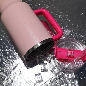 May include: A pink insulated tumbler with a bright pink handle and lid. The tumbler is partially filled with ice cubes, and water droplets are visible on the exterior. The lid is clear with a pink slider and a straw.