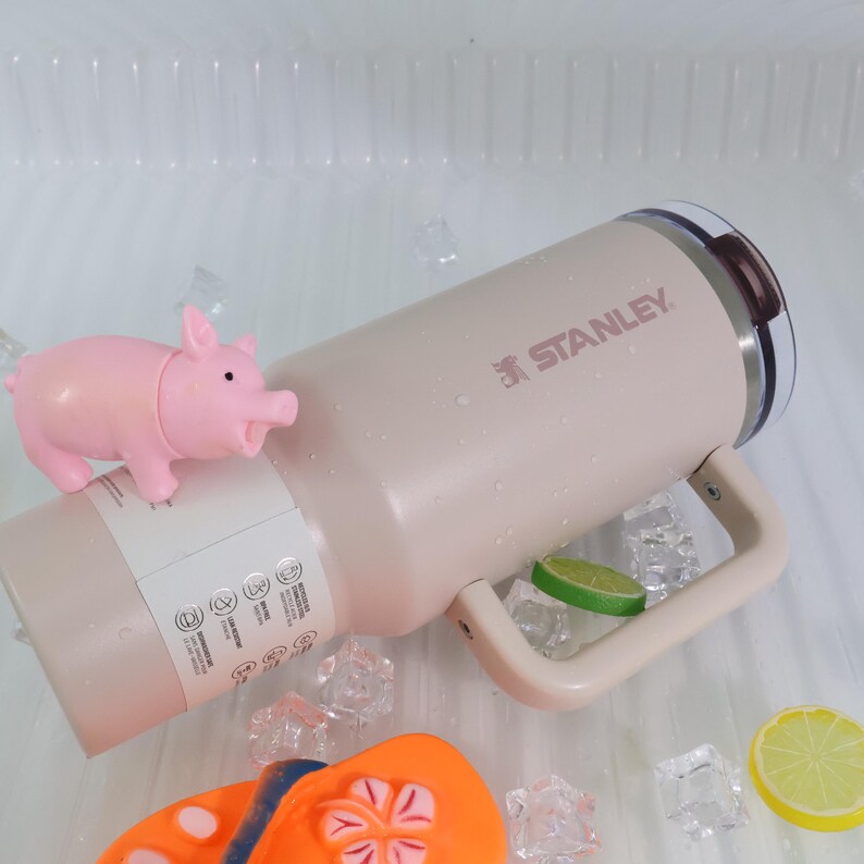 Peut inclure: Une tasse isotherme Stanley beige avec une poign&eacute;e, le nom de la marque imprim&eacute; en rose. Une petite figurine de cochon rose et des tongs orange et bleues sont &agrave; c&ocirc;t&eacute; de la tasse. La tasse est entour&eacute;e de gla&ccedil;ons et d'une tranche de citron vert et de citron.