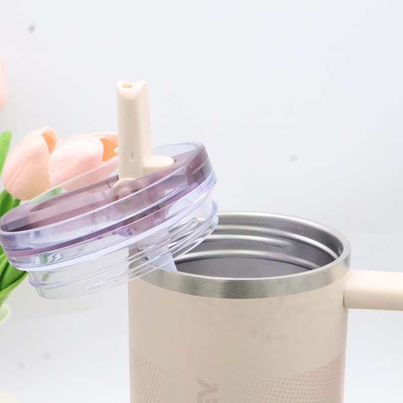 Puede incluir: Un vaso de acero inoxidable beige con una tapa transparente y una pajita beige. La tapa est&aacute; parcialmente abierta, revelando el interior del vaso. El vaso tiene un asa y un patr&oacute;n texturizado en la parte inferior.