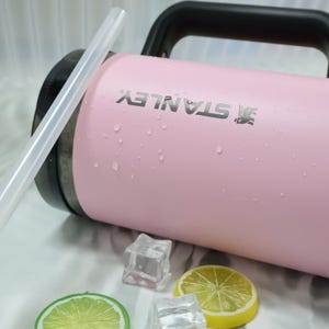 May include: A pink Stanley water bottle with a black handle and the Stanley logo. The bottle has a clear straw and is surrounded by ice cubes and lemon slices. The bottle appears to be wet with condensation.
