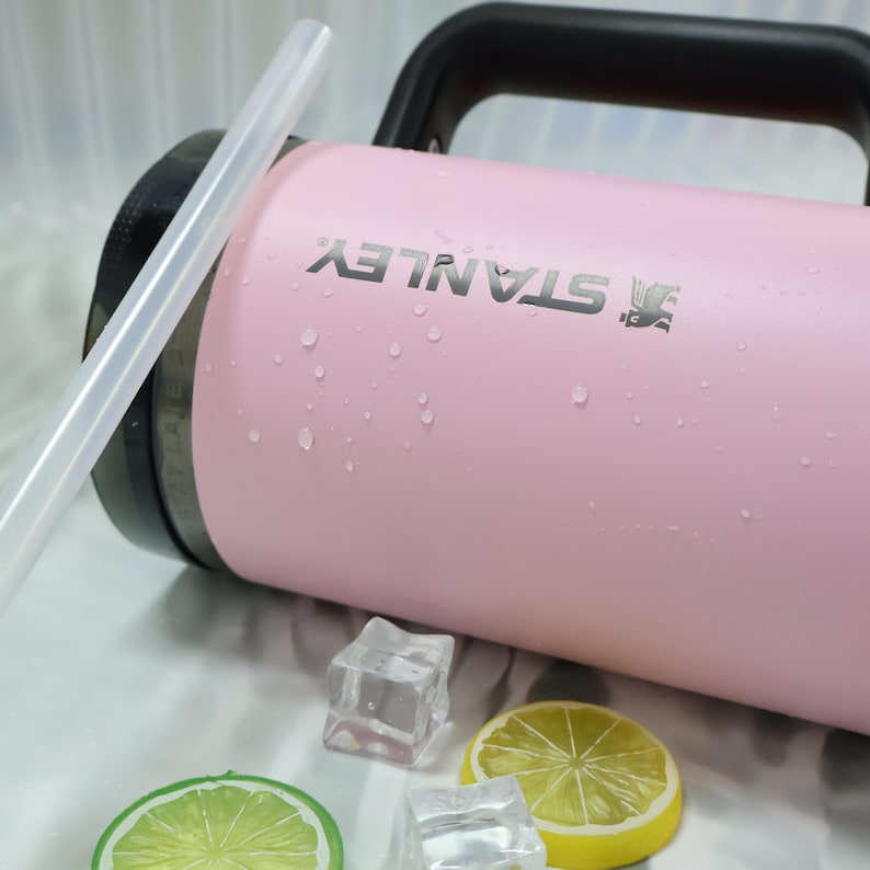 May include: A pink Stanley water bottle with a black handle and the Stanley logo. The bottle has a clear straw and is surrounded by ice cubes and lemon slices. The bottle appears to be wet with condensation.