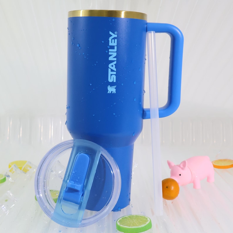May include: A blue Stanley tumbler with a gold rim, handle, and clear lid. The tumbler has the Stanley logo printed on the side. A clear straw and a small pink pig figurine are also present. The tumbler is approximately 9 inches tall.