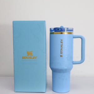 Pu&ograve; includere: Un bicchiere Stanley azzurro con manico e coperchio trasparente, accanto a una scatola abbinata. Il bicchiere ha il logo e il testo Stanley in oro. La scatola presenta anche il logo Stanley in oro.
