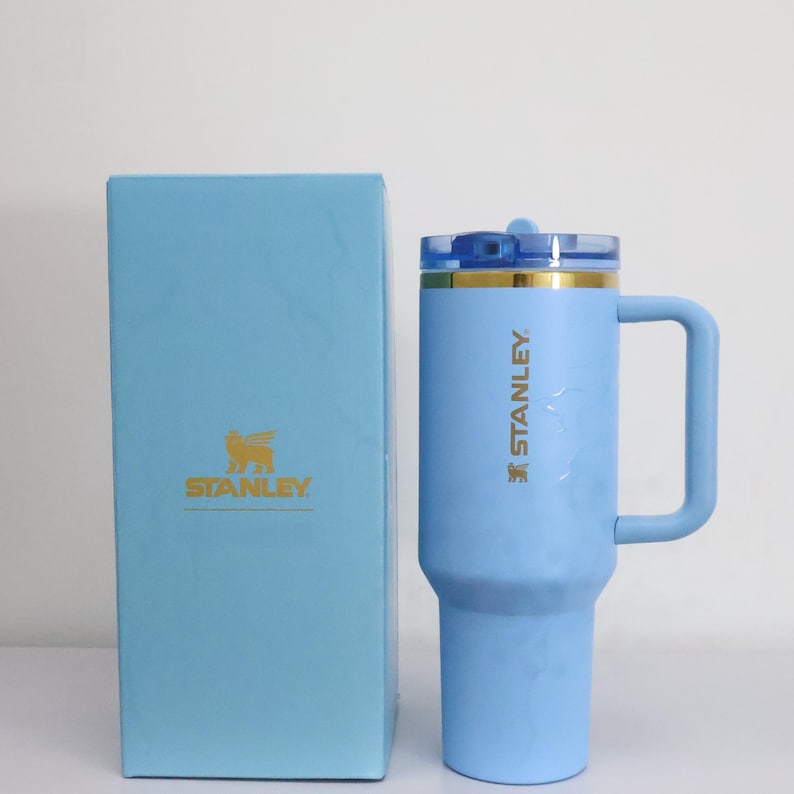 Pu&ograve; includere: Un bicchiere Stanley azzurro con manico e coperchio trasparente, accanto a una scatola abbinata. Il bicchiere ha il logo e il testo Stanley in oro. La scatola presenta anche il logo Stanley in oro.