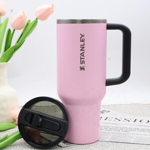 Pu&ograve; includere: Una tazza da viaggio Stanley rosa con manico e coperchio neri. La tazza ha il logo Stanley stampato sul lato. Il coperchio &egrave; nero e si trova accanto alla tazza. La tazza &egrave; su una superficie bianca.