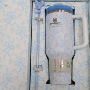 K&ouml;nnte beinhalten: Ein wei&szlig;-blauer Stanley Quencher-Becher mit Blumenmuster, passendem Strohhalm und Griff. Der Becher hat einen blauen Deckel und ist in einer Schachtel mit passendem Blumenmuster verpackt. Der Becher hat einen Durchmesser von 11 cm und ist 22 cm hoch.