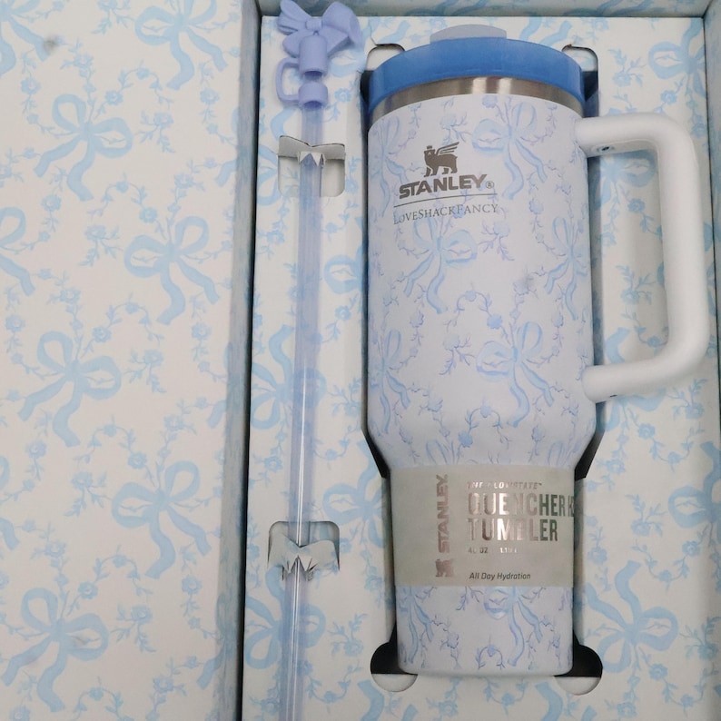 K&ouml;nnte beinhalten: Ein wei&szlig;-blauer Stanley Quencher-Becher mit Blumenmuster, passendem Strohhalm und Griff. Der Becher hat einen blauen Deckel und ist in einer Schachtel mit passendem Blumenmuster verpackt. Der Becher hat einen Durchmesser von 11 cm und ist 22 cm hoch.