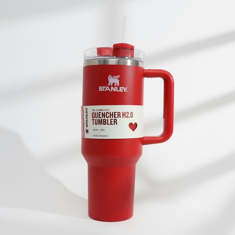 Może przedstawiać: Czerwony kubek Stanley Quencher H2.0 z uchwytem i słomką. Kubek ma białą etykietę z napisem "QUENCHER H2.0 TUMBLER" i symbolem serca. Kubek ma 1,18 L.