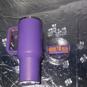 May include: A purple insulated tumbler with a handle and a lid. The handle and the lid's slider are a copper color. The tumbler is next to a clear straw and ice cubes. The tumbler is designed to keep drinks cold.