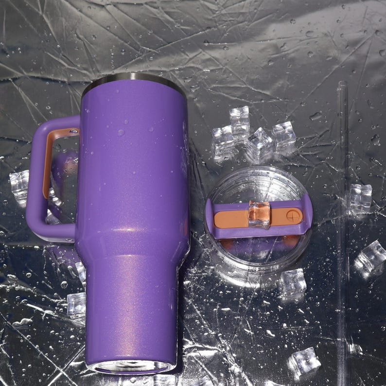 May include: A purple insulated tumbler with a handle and a lid. The handle and the lid's slider are a copper color. The tumbler is next to a clear straw and ice cubes. The tumbler is designed to keep drinks cold.