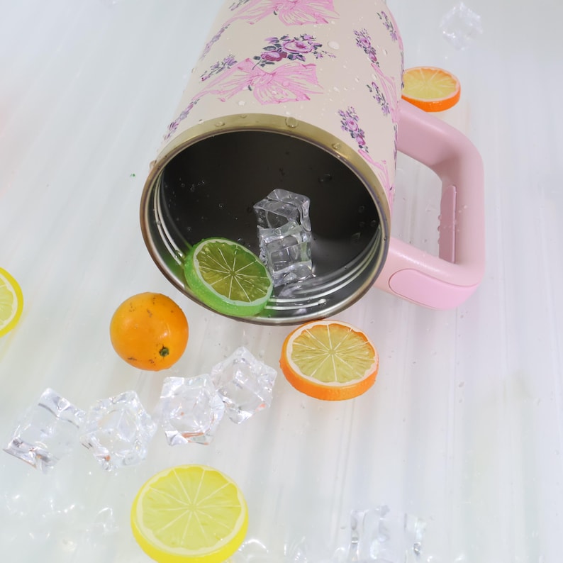 K&ouml;nnte beinhalten: Ein floral gemusterter Becher mit rosa Griff, der auf der Seite liegt. Im Inneren des Bechers befinden sich Eisw&uuml;rfel und eine Limettenscheibe. Verstreut sind Orangen- und Zitronenscheiben, mit mehr Eisw&uuml;rfeln.