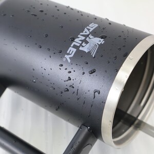 May include: A black Stanley thermos with the brand name and logo in white. The thermos has a silver rim and is covered in water droplets. The handle is black.