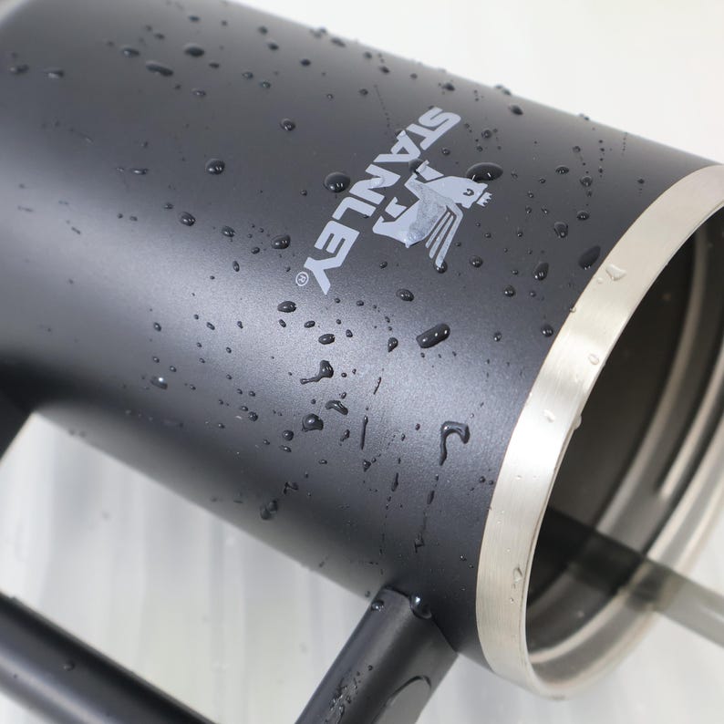 May include: A black Stanley thermos with the brand name and logo in white. The thermos has a silver rim and is covered in water droplets. The handle is black.