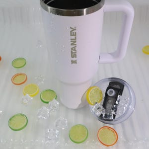 May include: White Stanley travel mug with a handle and a silver rim. The mug has the Stanley logo printed on the side. The lid is clear with a black mechanism. The mug is surrounded by ice cubes and lemon slices.