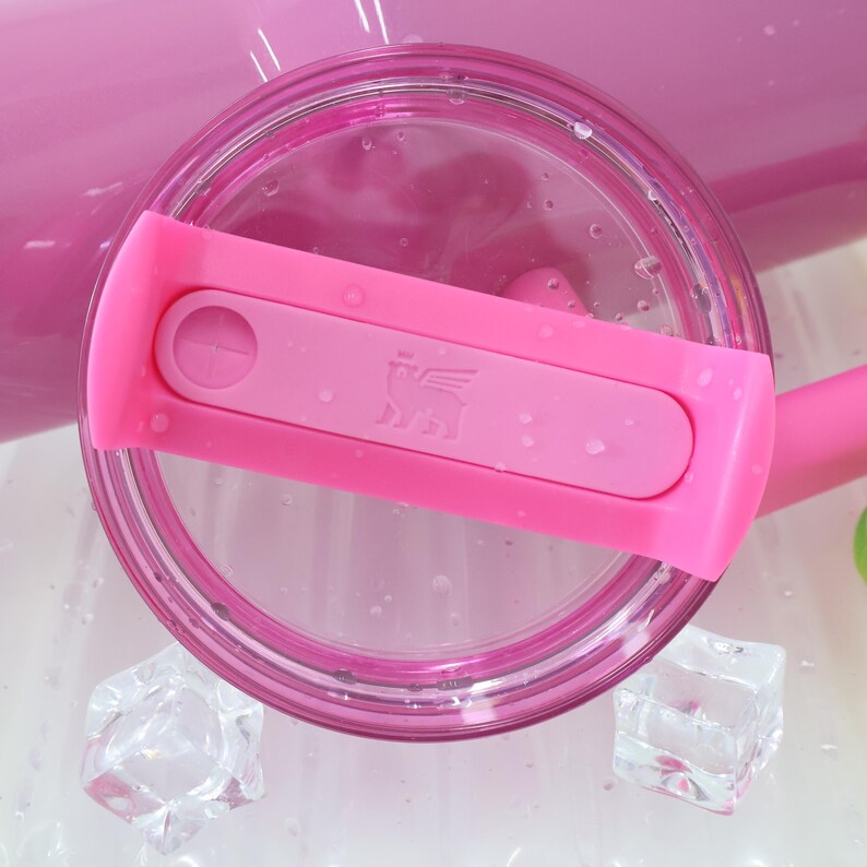 Op de afbeelding: Een close-up van een roze tumblerdeksel met een roze schuif en een doorzichtig deksel. De schuif heeft een ronde knop en een logo van een gevleugeld dier. Het deksel is omgeven door waterdruppels en ijsblokjes.