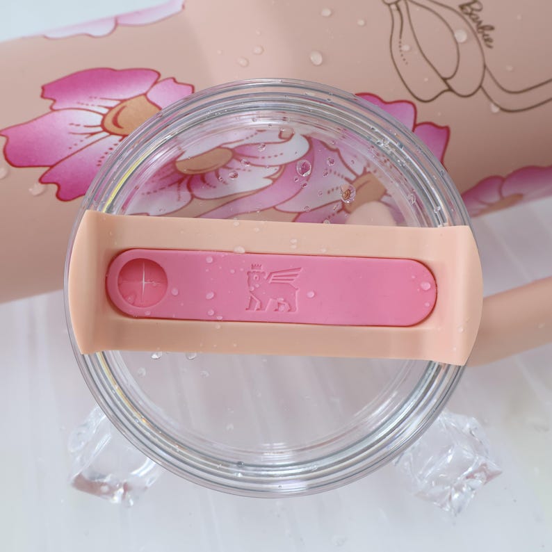 K&ouml;nnte beinhalten: Ein klarer, kreisf&ouml;rmiger Becherdeckel mit einem rosa Schieber und einem pfirsichfarbenen Band. Der Schieber hat ein Logo und einen kleinen Kreis. Der Hintergrund zeigt ein Blumendesign und das Wort "Barbie".