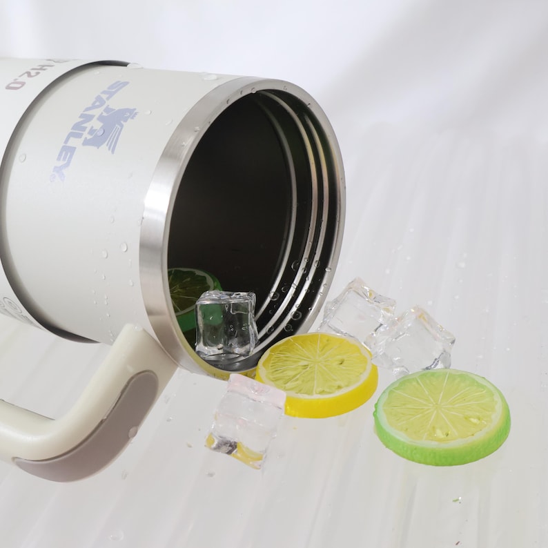 May include: A white Stanley H2.0 insulated tumbler with a silver rim and handle, shown with ice cubes, lemon and lime slices. The tumbler is partially tipped, with water droplets visible on the surface. The Stanley logo is printed on the side.
