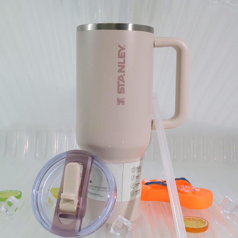 Peut inclure: Un mug Stanley rose clair avec un bord et une poign&eacute;e argent&eacute;s. Le mug porte le logo Stanley imprim&eacute; sur le c&ocirc;t&eacute;. Un couvercle transparent et une paille sont &agrave; c&ocirc;t&eacute; du mug. Le mug est pos&eacute; sur une surface blanche avec des gla&ccedil;ons et des tranches de fruits.