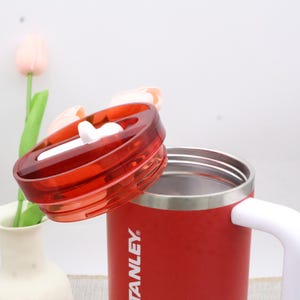 Pu&ograve; includere: Una tazza da viaggio Stanley rossa con manico bianco e coperchio rosso trasparente. Il coperchio &egrave; parzialmente aperto, rivelando l'interno in acciaio inossidabile. La tazza &egrave; su una superficie bianca con un vaso e fiori sullo sfondo.