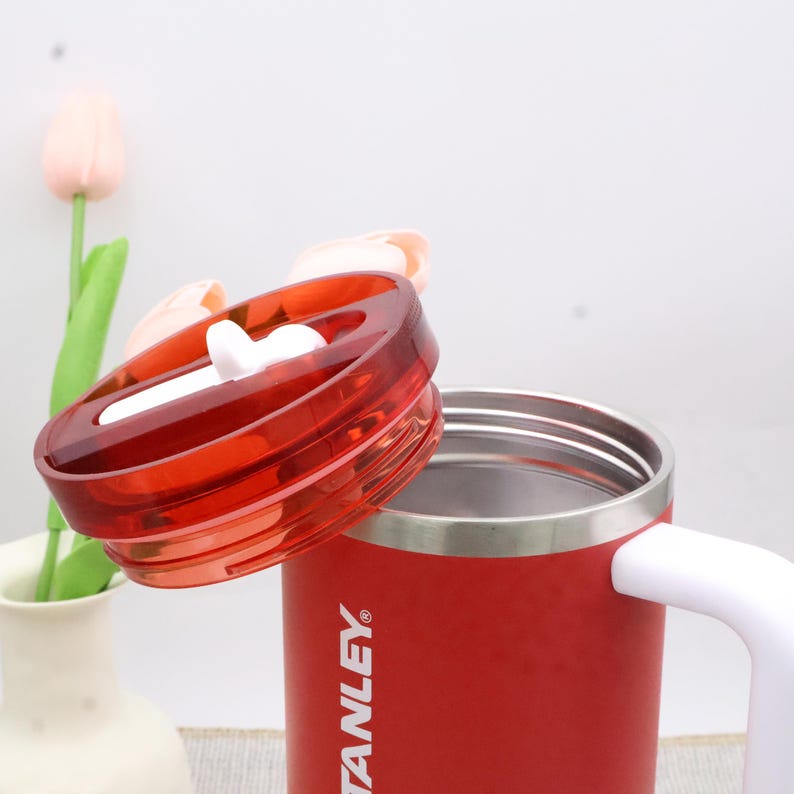 Pu&ograve; includere: Una tazza da viaggio Stanley rossa con manico bianco e coperchio rosso trasparente. Il coperchio &egrave; parzialmente aperto, rivelando l'interno in acciaio inossidabile. La tazza &egrave; su una superficie bianca con un vaso e fiori sullo sfondo.