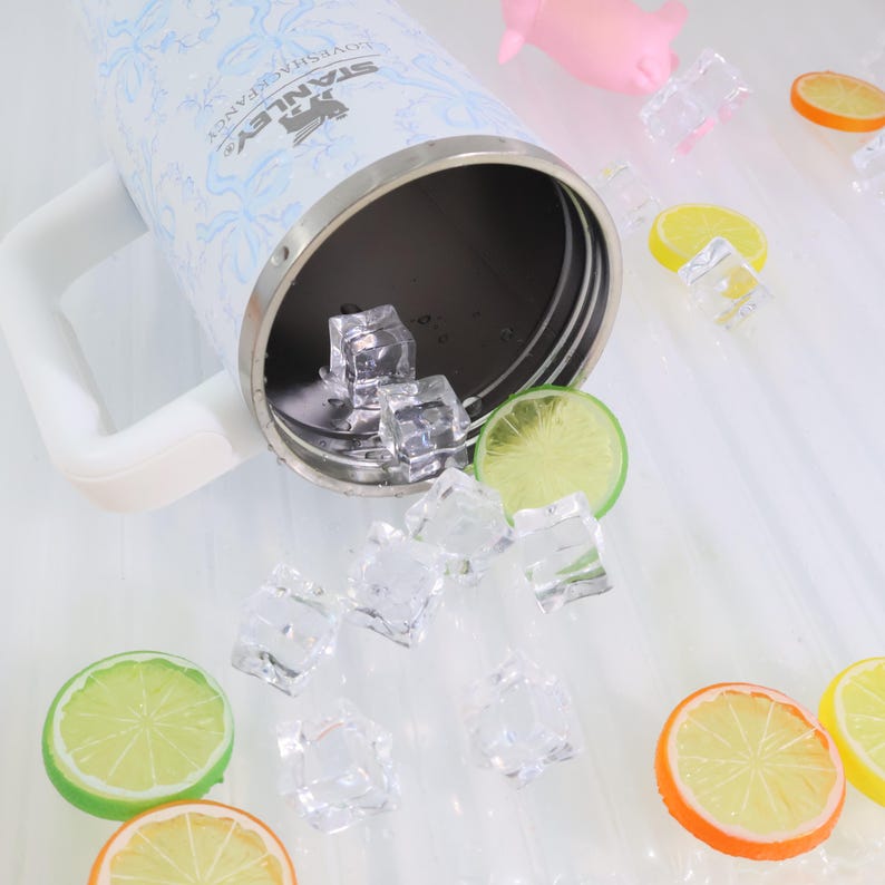 K&ouml;nnte beinhalten: Ein Stanley-Becher aus Edelstahl mit wei&szlig;em Griff und hellblauem Blumenmuster. Der offene Becher enth&auml;lt Eisw&uuml;rfel. Zitronen- und Limettenscheiben sind zusammen mit weiteren Eisw&uuml;rfeln um den Becher verstreut, was eine erfrischende &Auml;sthetik erzeugt.