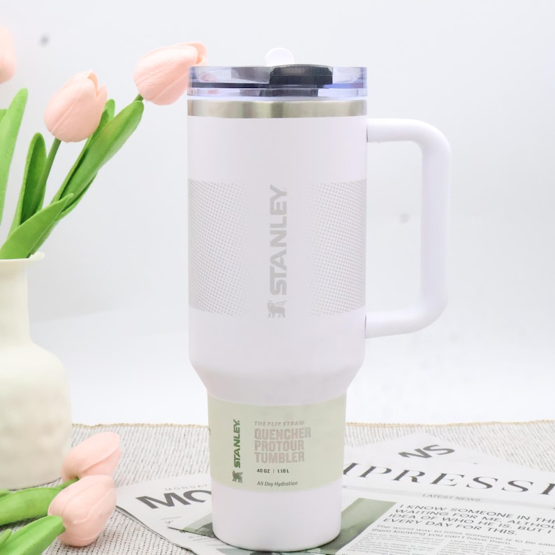 Puede incluir: Vaso Stanley Quencher Protour blanco con asa y tapa transparente. El vaso tiene el logotipo de Stanley y texto en el lateral. Dise&ntilde;ado para la hidrataci&oacute;n durante todo el d&iacute;a.