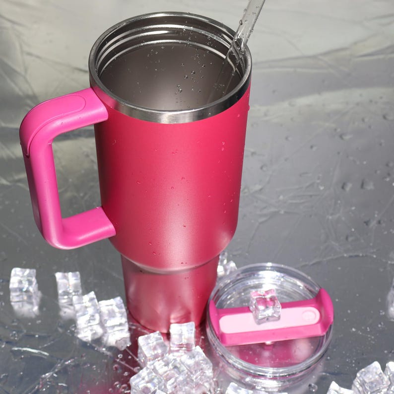 Peut inclure: Un grand gobelet isotherme magenta vif, avec une poign&eacute;e et un couvercle assortis. Le bord en acier inoxydable et un couvercle transparent avec un curseur rose sont visibles. Le gobelet est pr&eacute;sent&eacute; avec des gla&ccedil;ons et une paille.