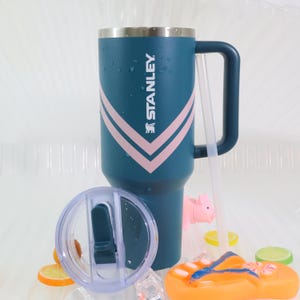 K&ouml;nnte beinhalten: Ein tealfarbener Stanley-Becher mit Griff und rosa Chevron-Design. Der Edelstahlbecher hat das Stanley-Logo und einen transparenten Deckel. Ein Strohhalm und dekorative Elemente befinden sich im Vordergrund.