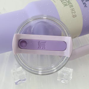 Może przedstawiać: Zbliżenie lawendowego kubka Stanley Quencher H2.0 FlowState z przezroczystą pokrywką. Pokrywka posiada lawendowy suwak z logo i przezroczystą plastikową obręczą. Dwie kostki lodu na pierwszym planie.