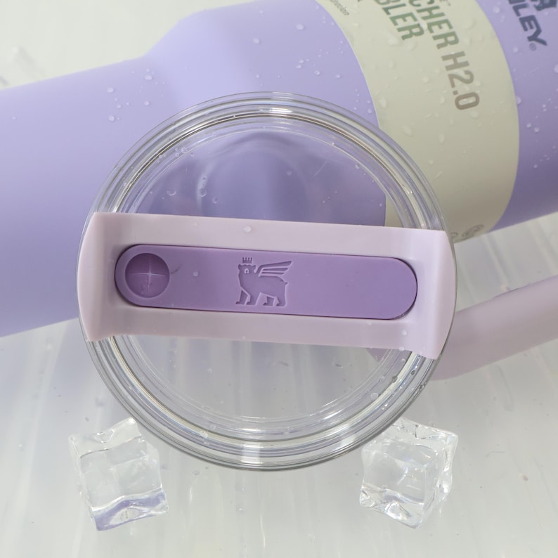 Może przedstawiać: Zbliżenie lawendowego kubka Stanley Quencher H2.0 FlowState z przezroczystą pokrywką. Pokrywka posiada lawendowy suwak z logo i przezroczystą plastikową obręczą. Dwie kostki lodu na pierwszym planie.