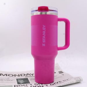 Puede incluir: Un termo de viaje Stanley rosa brillante con asa. El termo tiene una tapa transparente con la parte superior rosa y el logotipo de Stanley. El termo est&aacute; sobre un peri&oacute;dico. El termo mide unos 23 cm de alto.