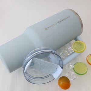 Puede incluir: Un vaso Stanley Starbucks azul claro con tapa transparente y asa, con el nombre de la marca impreso en el lateral. El vaso est&aacute; junto a rodajas de lim&oacute;n y lima, cubitos de hielo y una peque&ntilde;a naranja.